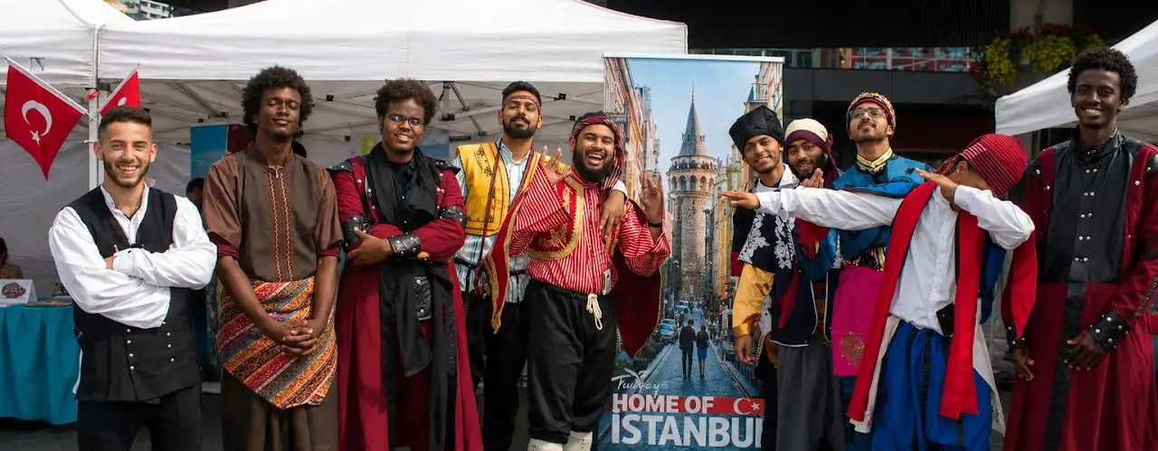 A group of performers in traditional Anatolian costumes pose in front of a Türkiye tourism banner showing Galata Tower.