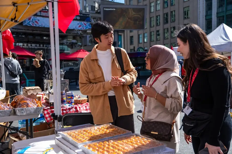 Visitors chat at a vendor stand laid out with trays of baklava.