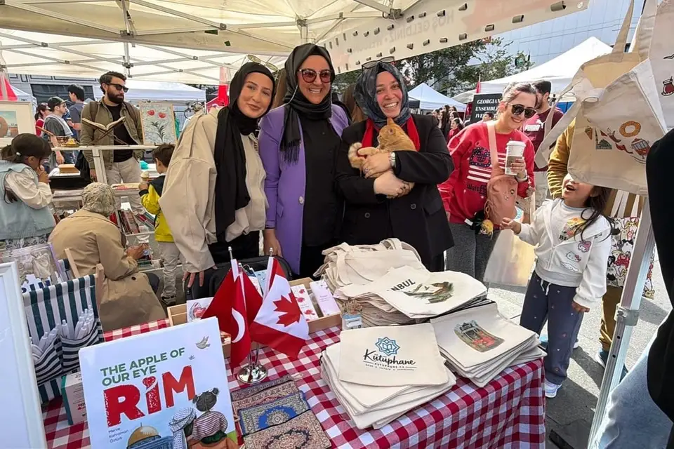 A stand with books, printed tote bags and small Turkish and Canadian flags, visitors browsing behind.