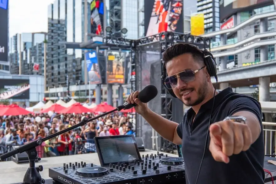 The DJ points at the camera from the stage, the square full of people behind him.