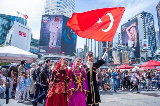 A Turkish flag waves above performers in traditional dress crossing the square, billboards behind.