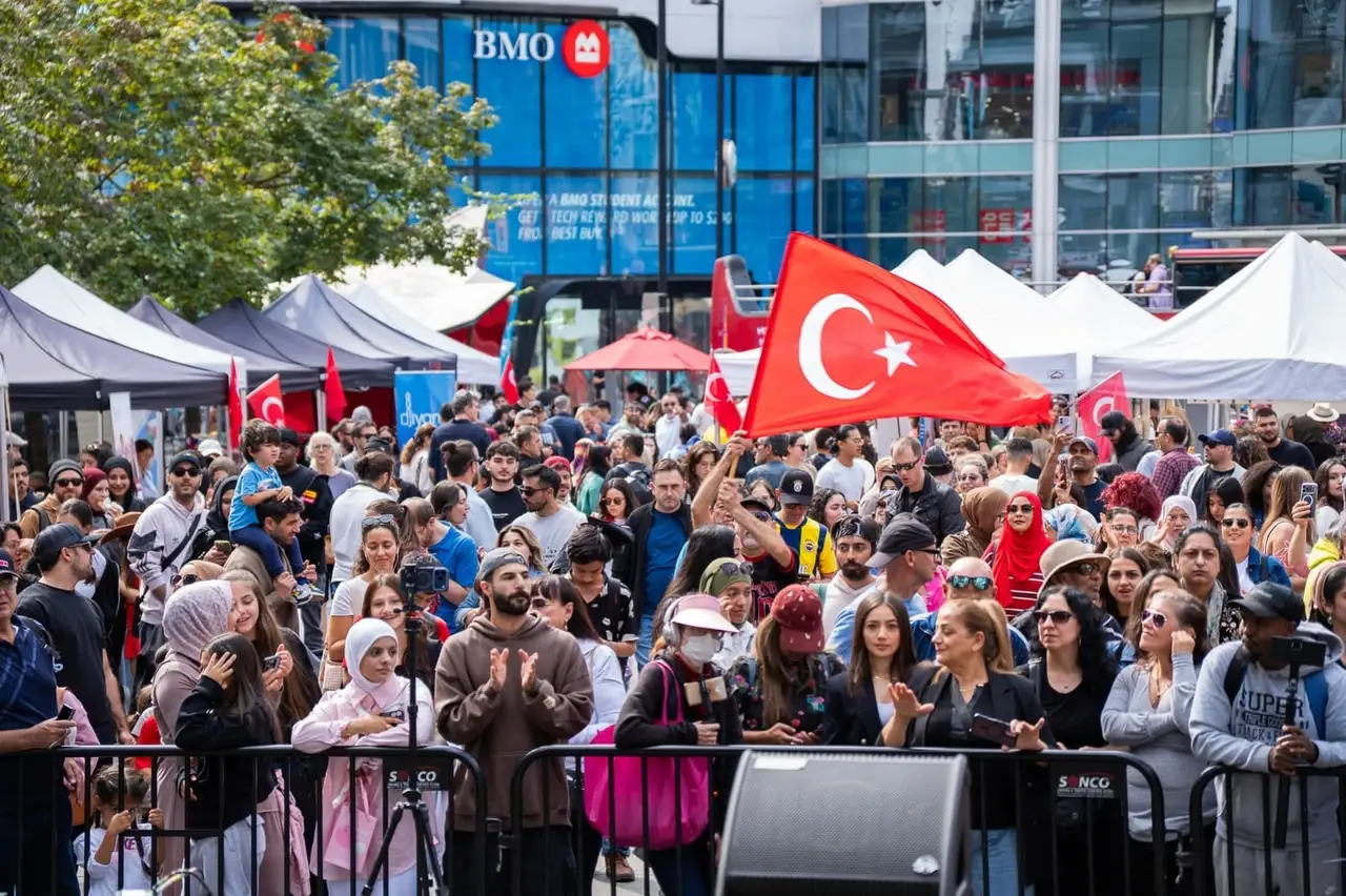 A large Turkish flag rises above the crowd in front of the stage, white market tents behind.