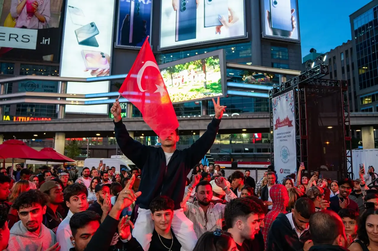 A young man raised above the evening crowd waves a Turkish flag, the square’s screens lit behind him.