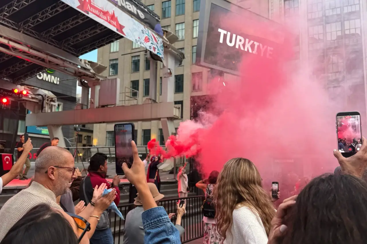 Red smoke drifts over the crowd while visitors film on their phones; a TÜRKİYE sign glows on the screen behind.