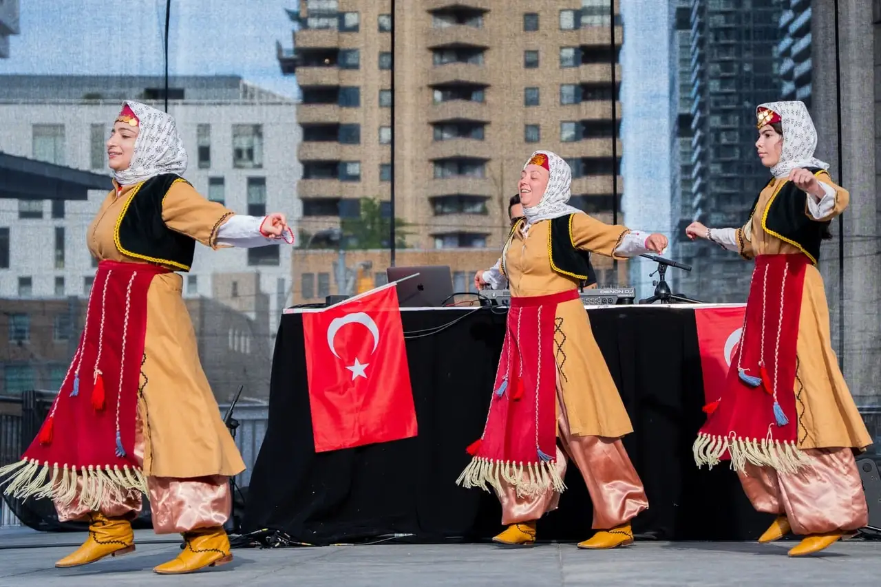 Three dancers in red-and-gold costumes perform on stage in front of a Turkish flag.