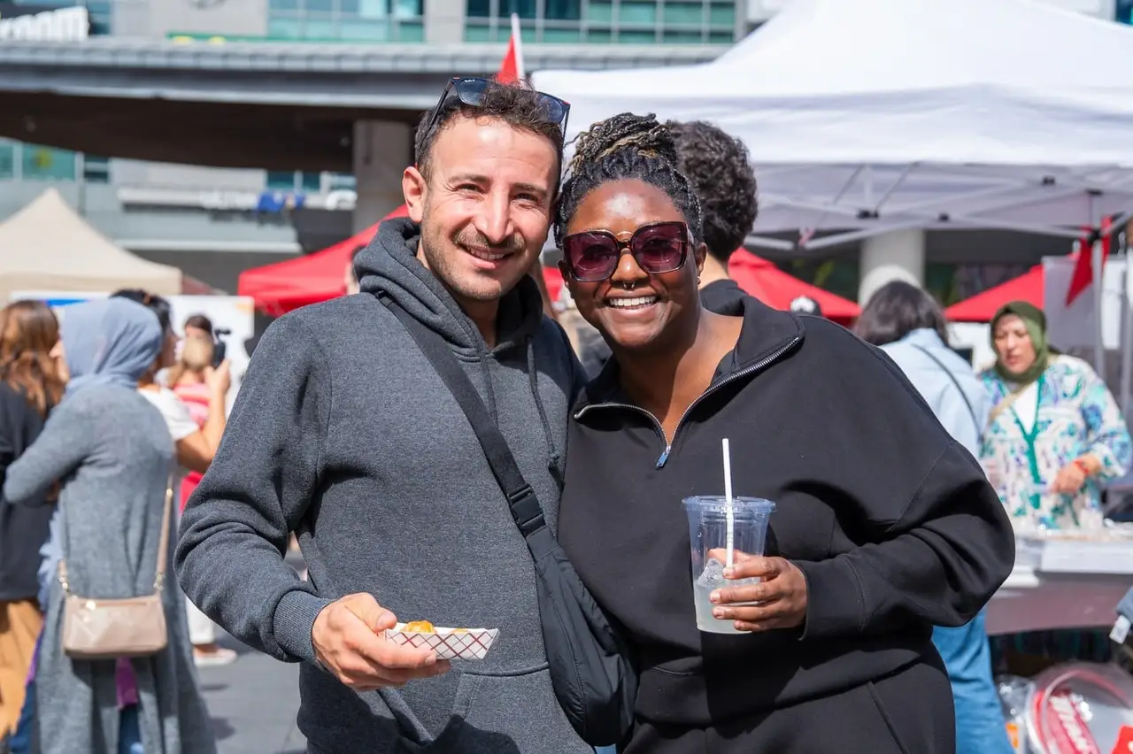 Two visitors smile for the camera, a drink in hand, the market tents behind them.