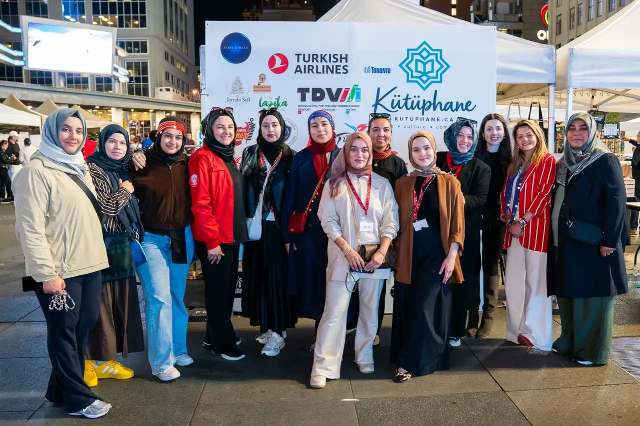 A group of women in red scarves stands in front of the festival’s sponsor wall in the evening.