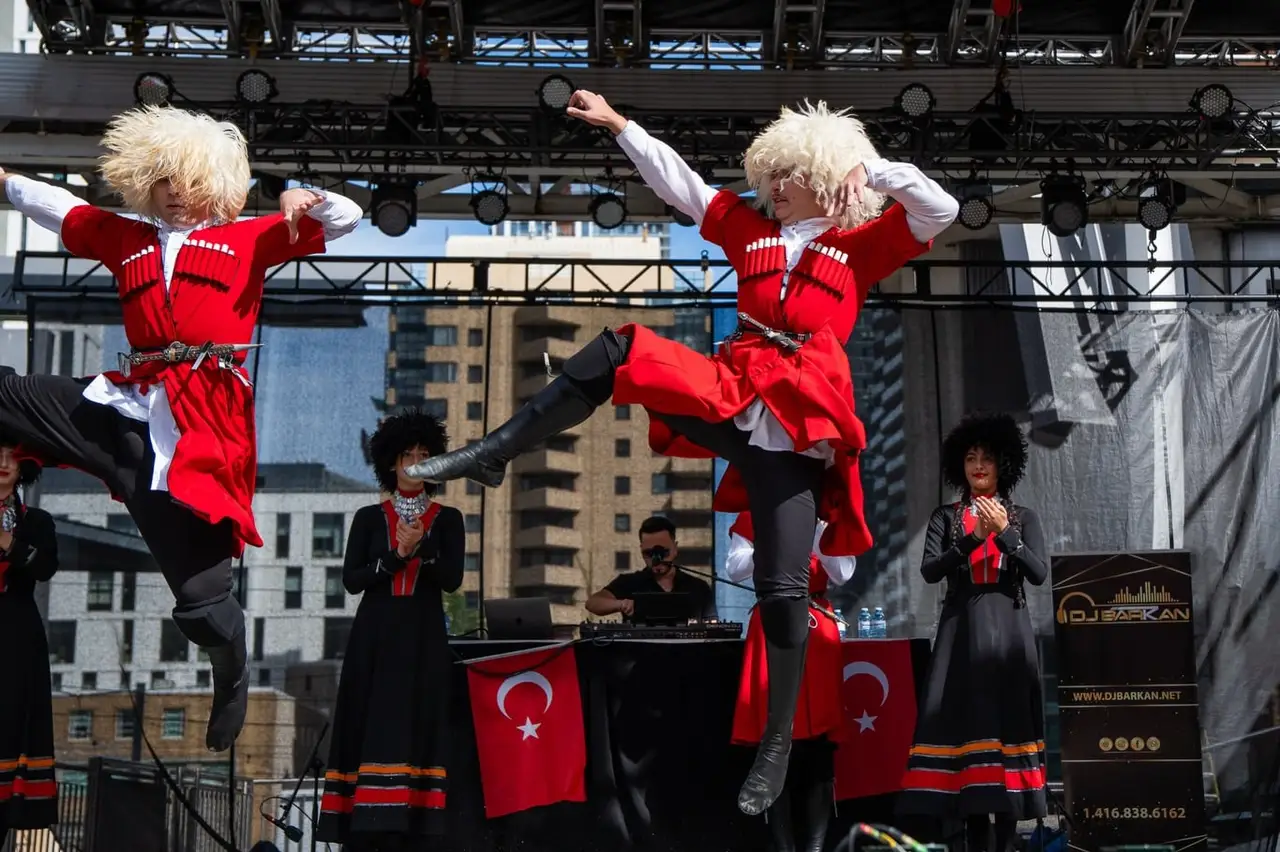 Two dancers in red costumes and white fur hats leap across the main stage while dancers behind them clap; Turkish flags hang from the DJ table.