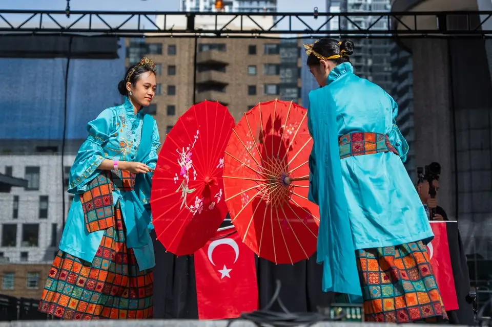 Two performers in turquoise costumes hold red parasols on stage, a Turkish flag draped between them.