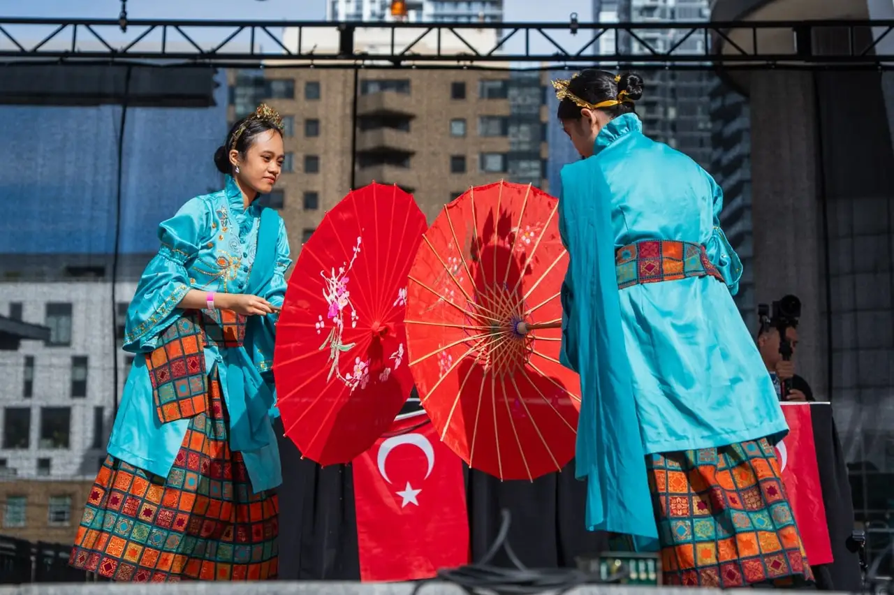 Two performers in turquoise costumes hold red parasols on stage, a Turkish flag draped between them.