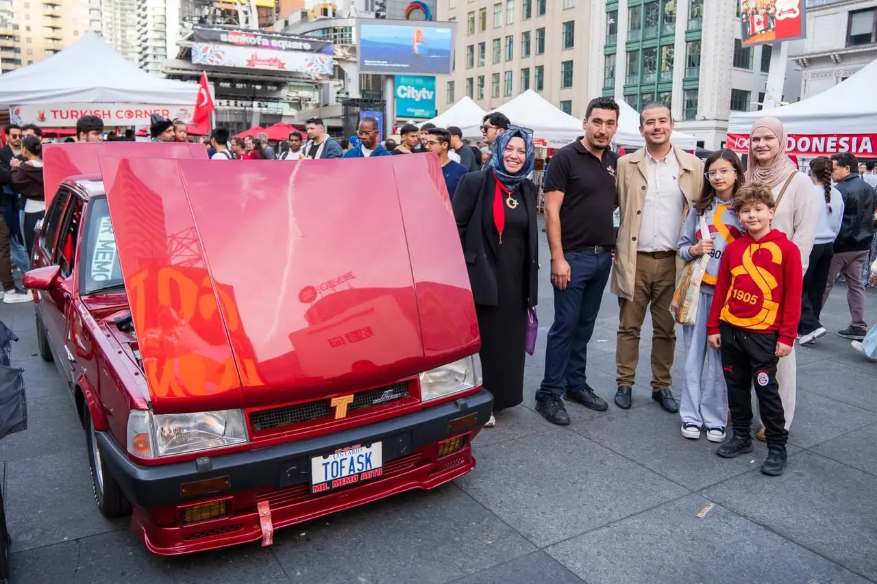 A classic red Tofaş with its bonnet up, visitors gathered around it on the square.