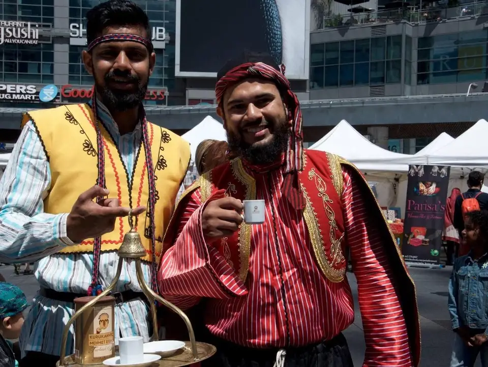 Two men in Ottoman-style costumes, one holding a brass coffee pot, pose with a cup of Turkish coffee.