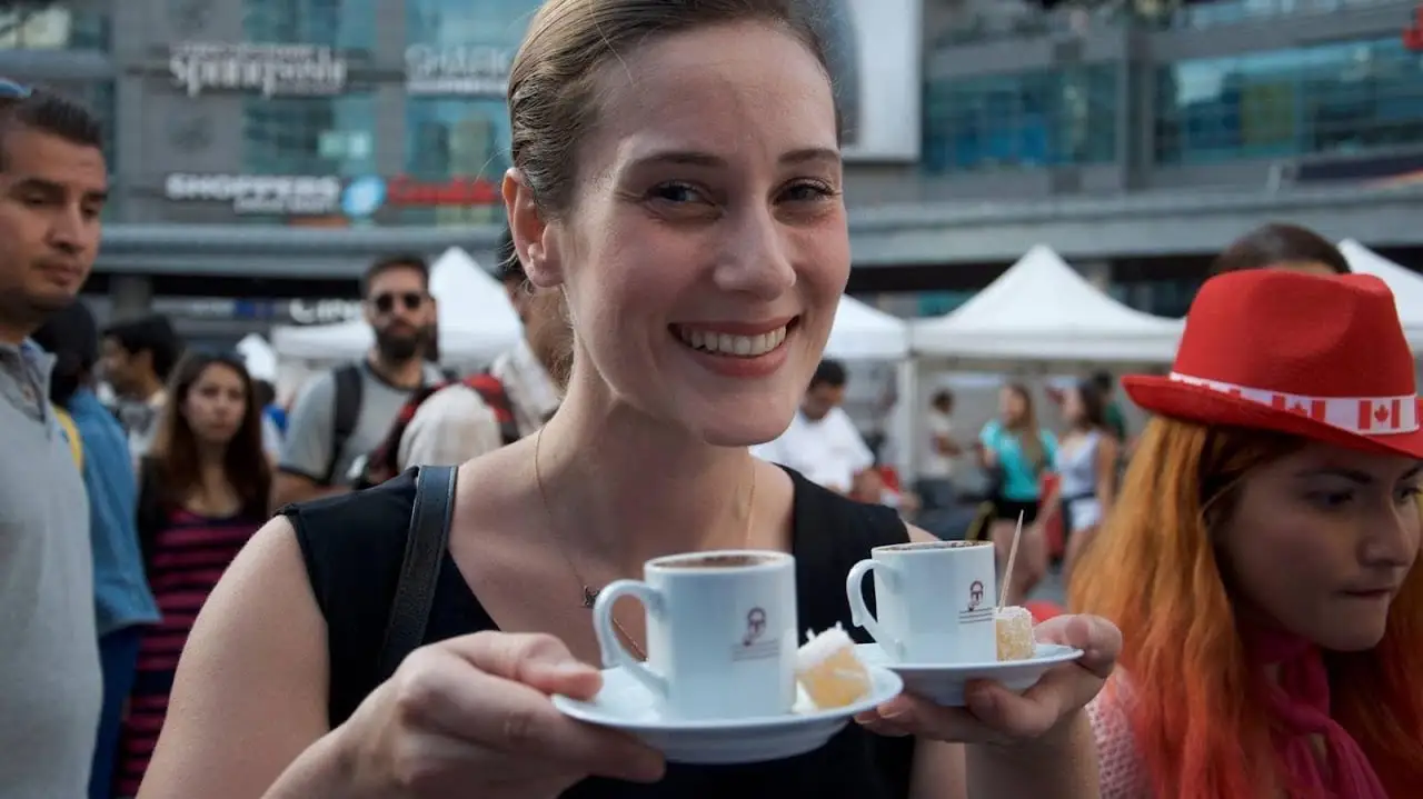 A smiling visitor carries two cups of Turkish coffee on a saucer through the crowd.