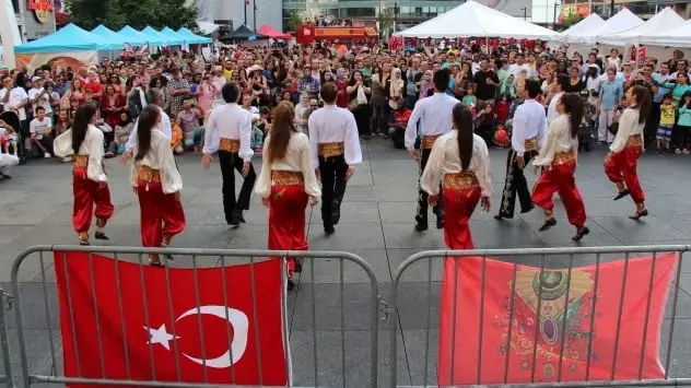 Folk dancers in red and white perform on the square, flags hung on the barrier in front.