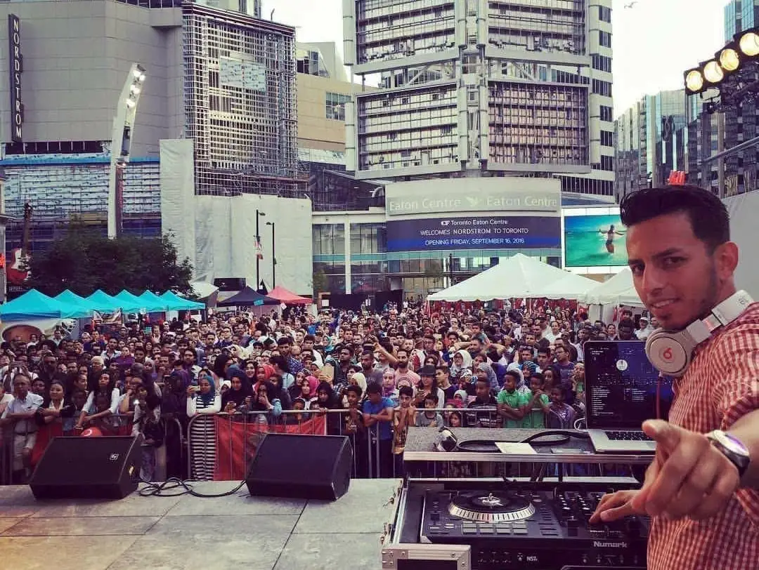 A DJ points at the camera from the stage, the square full of people behind him.